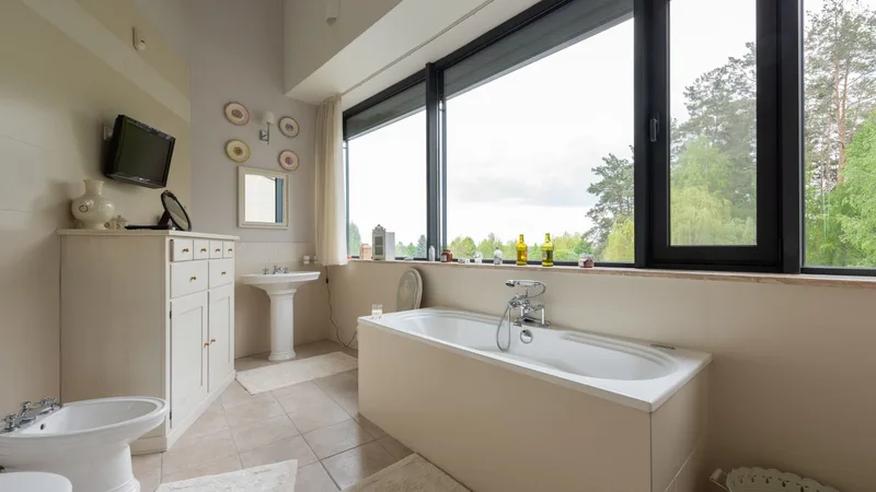 Büyük pencereli, yeşilliğe bakan ferah bir banyo, küvet ve lavabo
