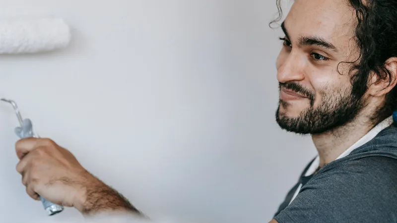 Beyaz tişörtlü bir kişinin rulo fırçayla iç mekan duvarını boyarken gülümsemesi